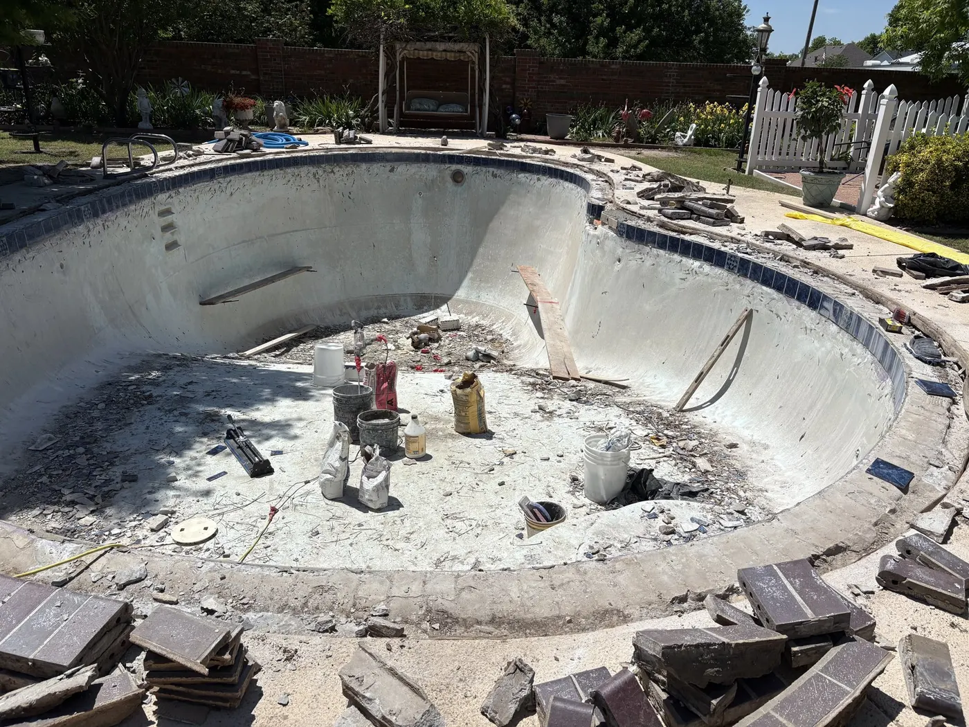 Drained kidney-shaped pool with old tile chipped off and debris on the floor before renovation
