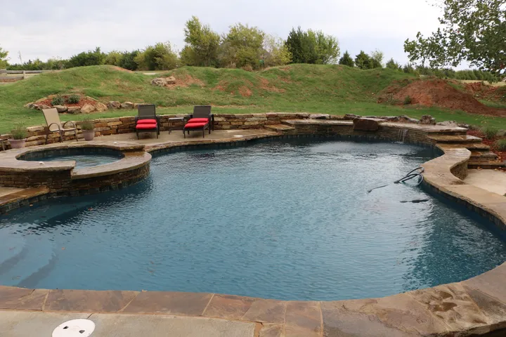 Contemporary freeform pool with stone walls, concrete deck, and red lounge chairs in landscaped setting