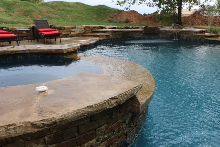 Landscape shot of curved freeform pool with stone walls, red lounge chairs, and sloped landscaping