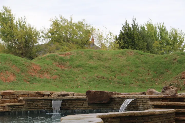 Stone-walled freeform pool with waterfall feature and grassy hillside backdrop