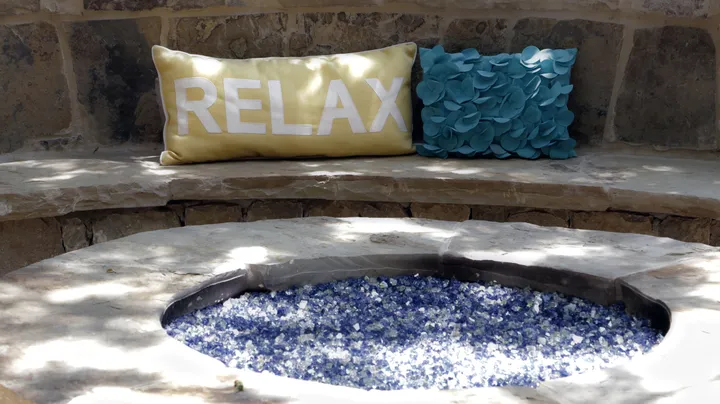Circular gas fire pit with blue fire glass and RELAX pillow in upscale stone seating area