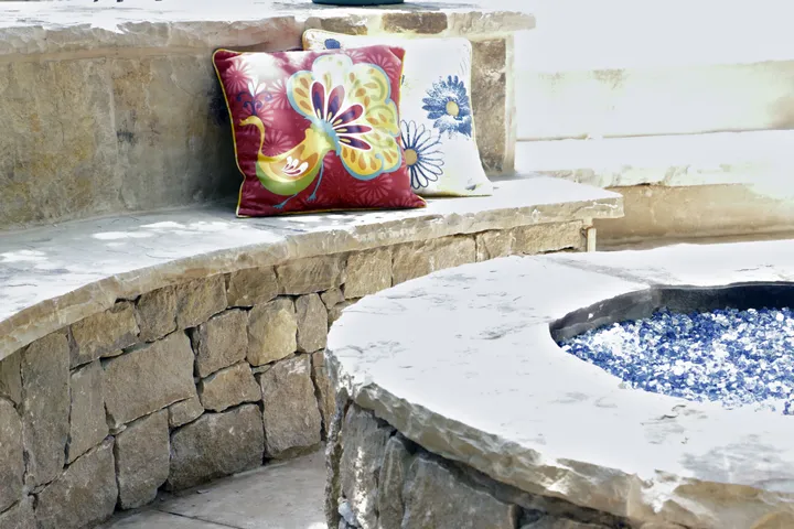 Circular stone fire pit with blue fire glass and decorative cushions on stone bench seating